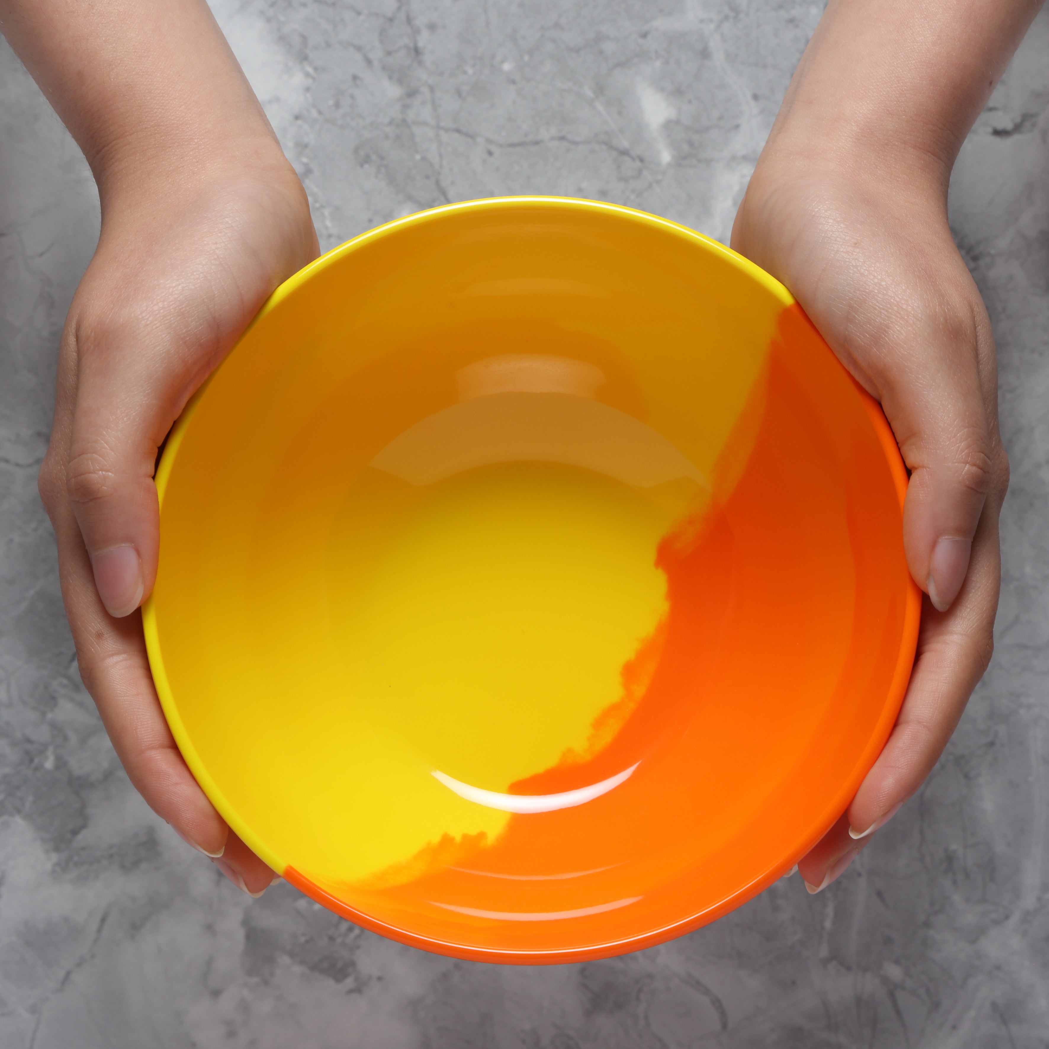 Two hands holding a Prue Leith melamine yellow and orange bowl against a gray background