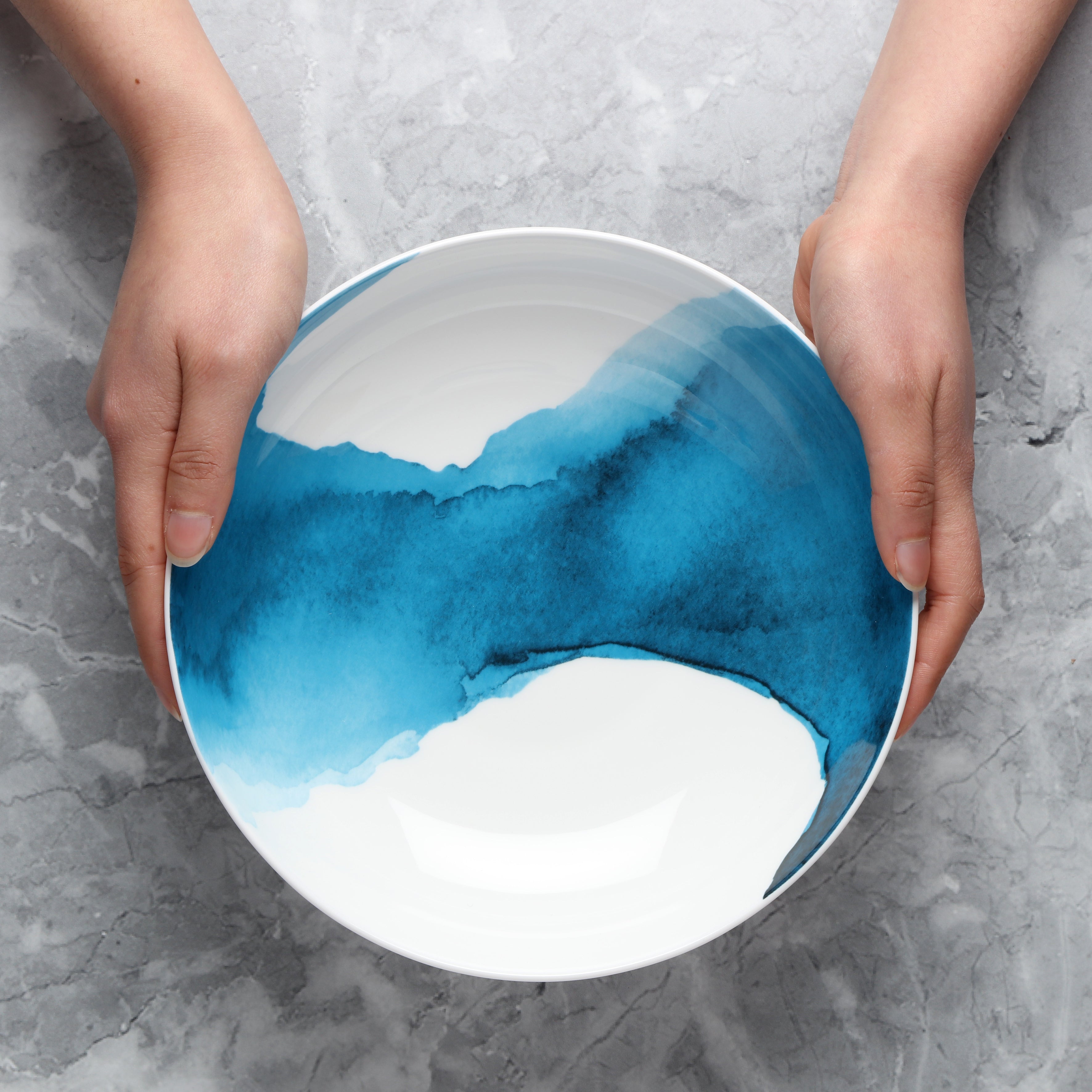Person holding a ceramic supper bowl with blue and white design on a gray surface overhead