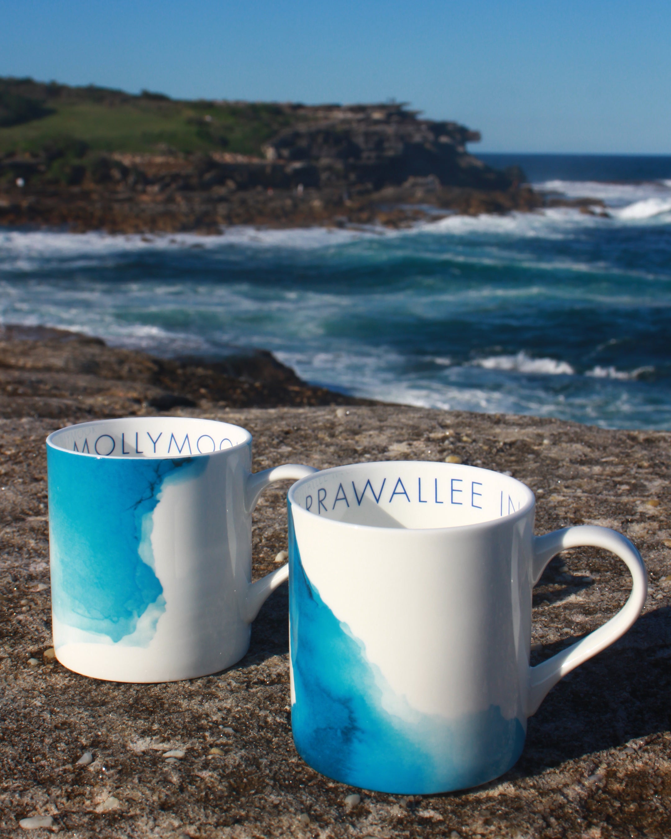 Two mugs with blue and white watercolour designs on a rock with ocean waves in the background.
