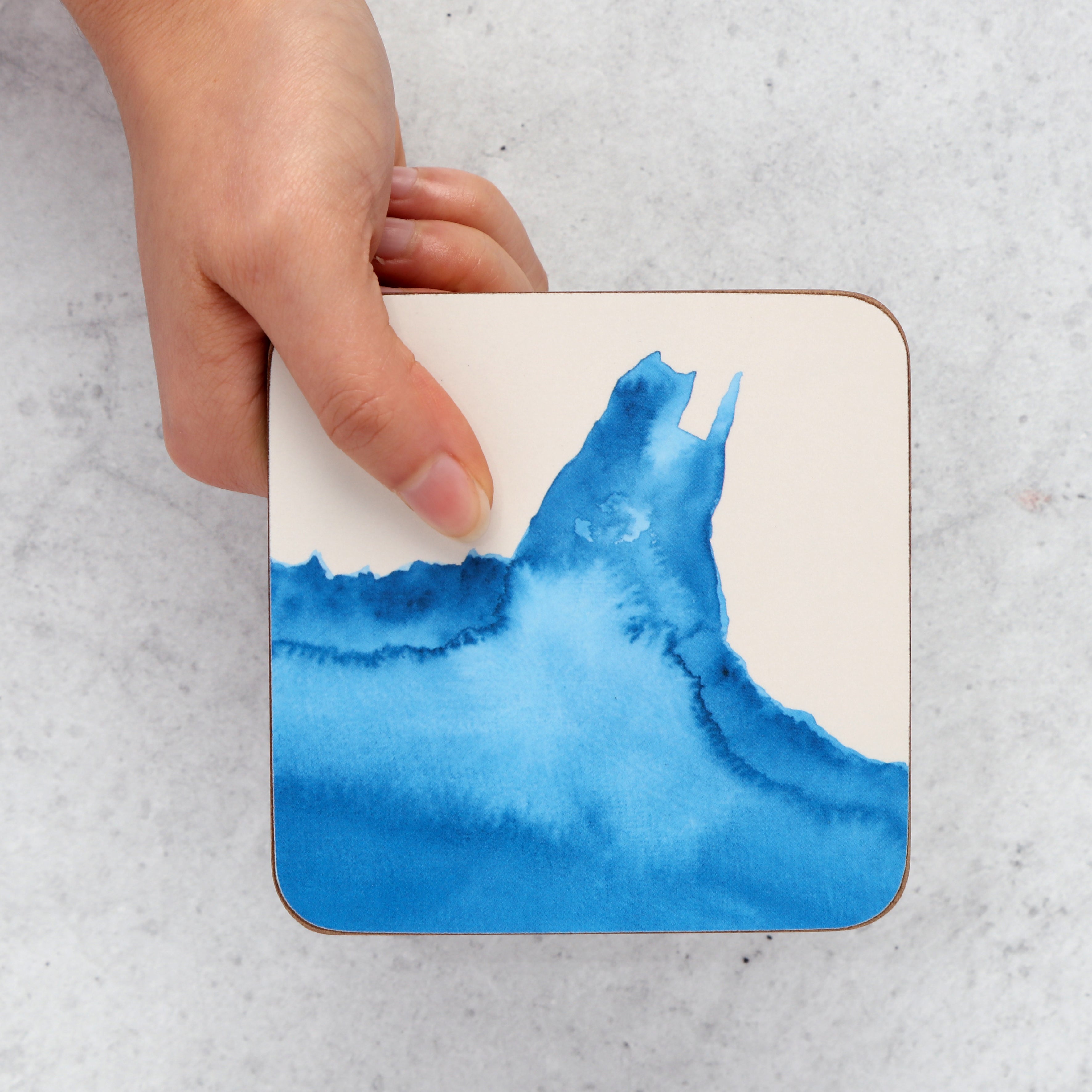 Coaster with blue watercolour design held by a hand on a textured surface