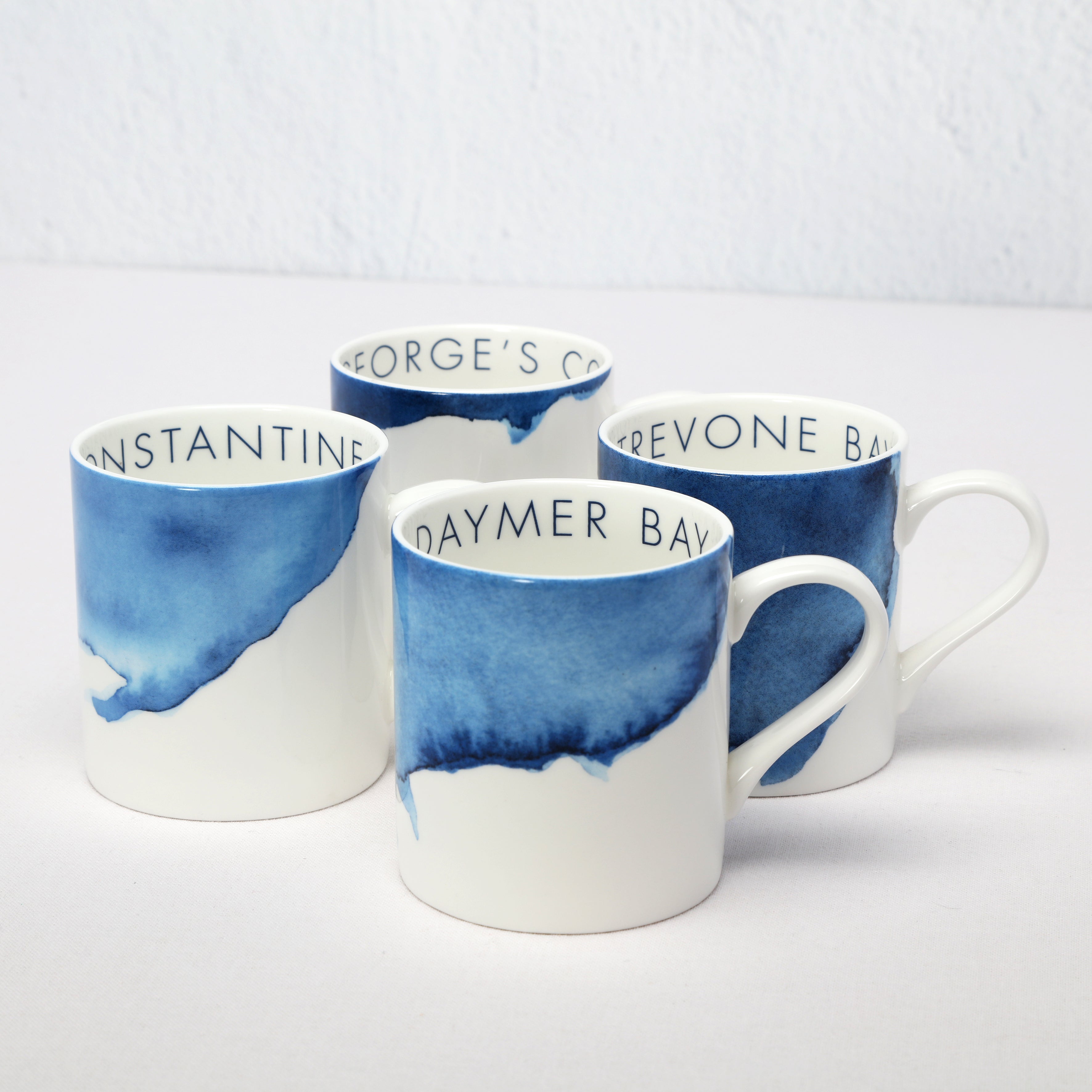 Four bone china mugs with blue watercolor designs and text on a white background