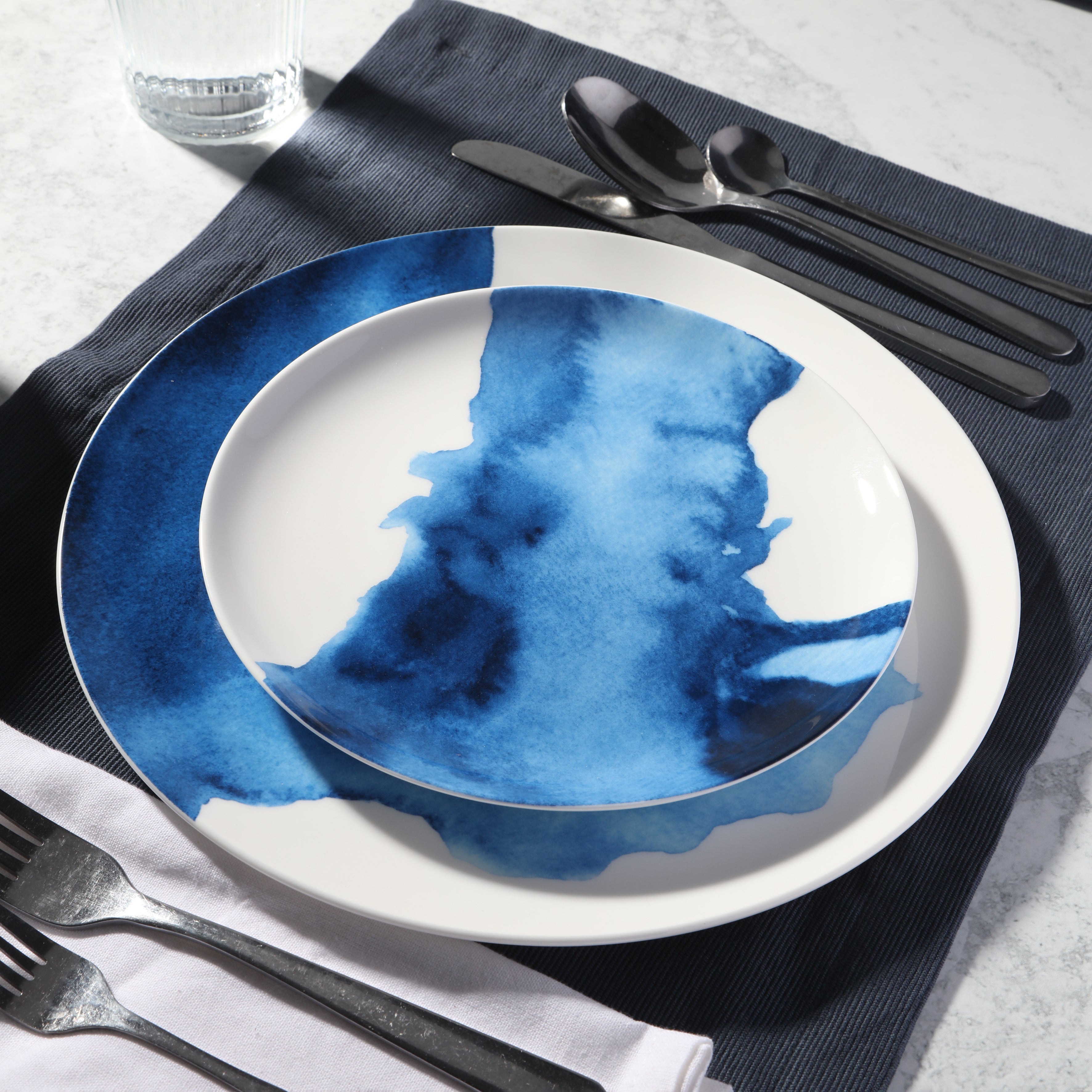 White plate with blue watercolor design on a table setting with silverware and napkin.