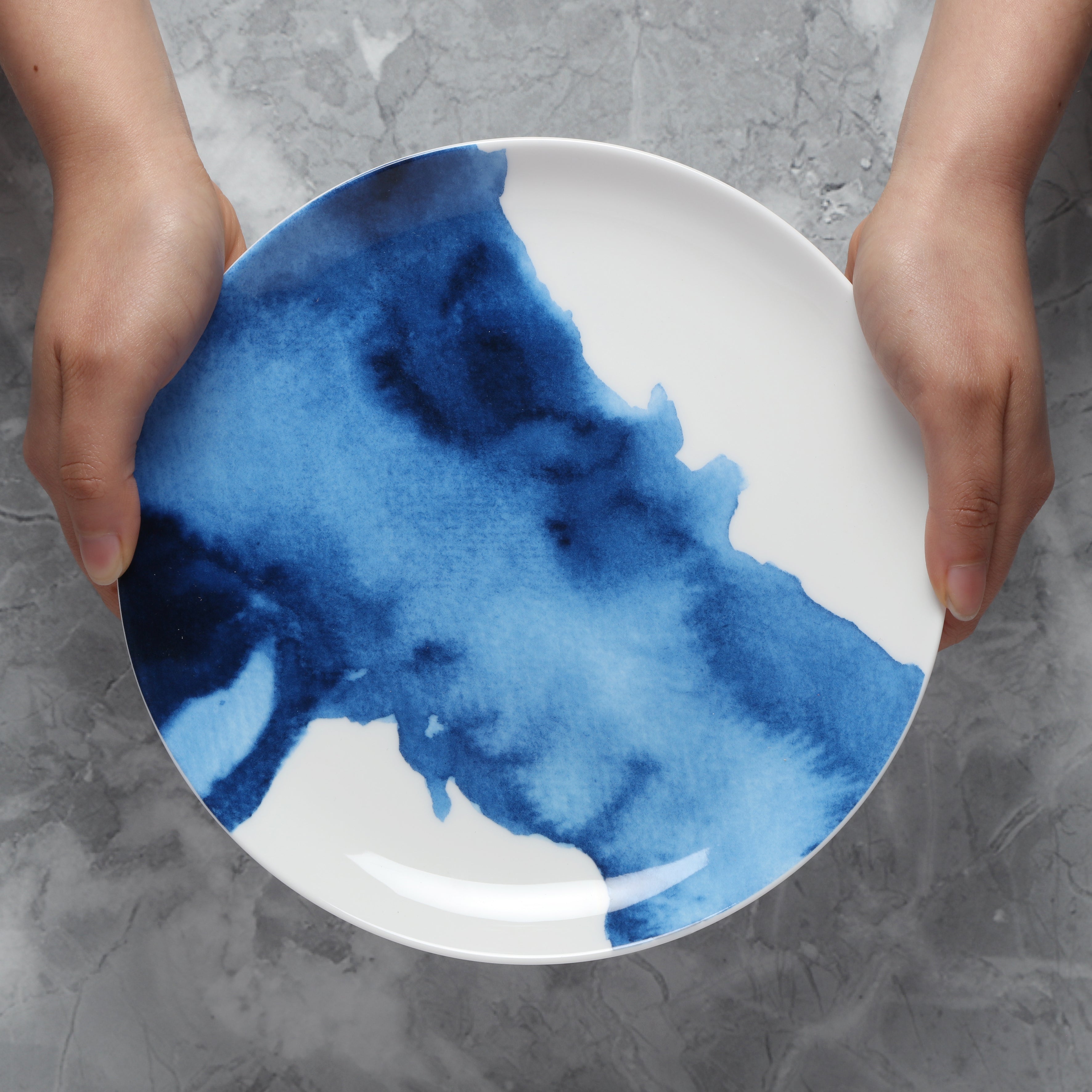 Person holding a white plate with blue watercolour design on a gray surface