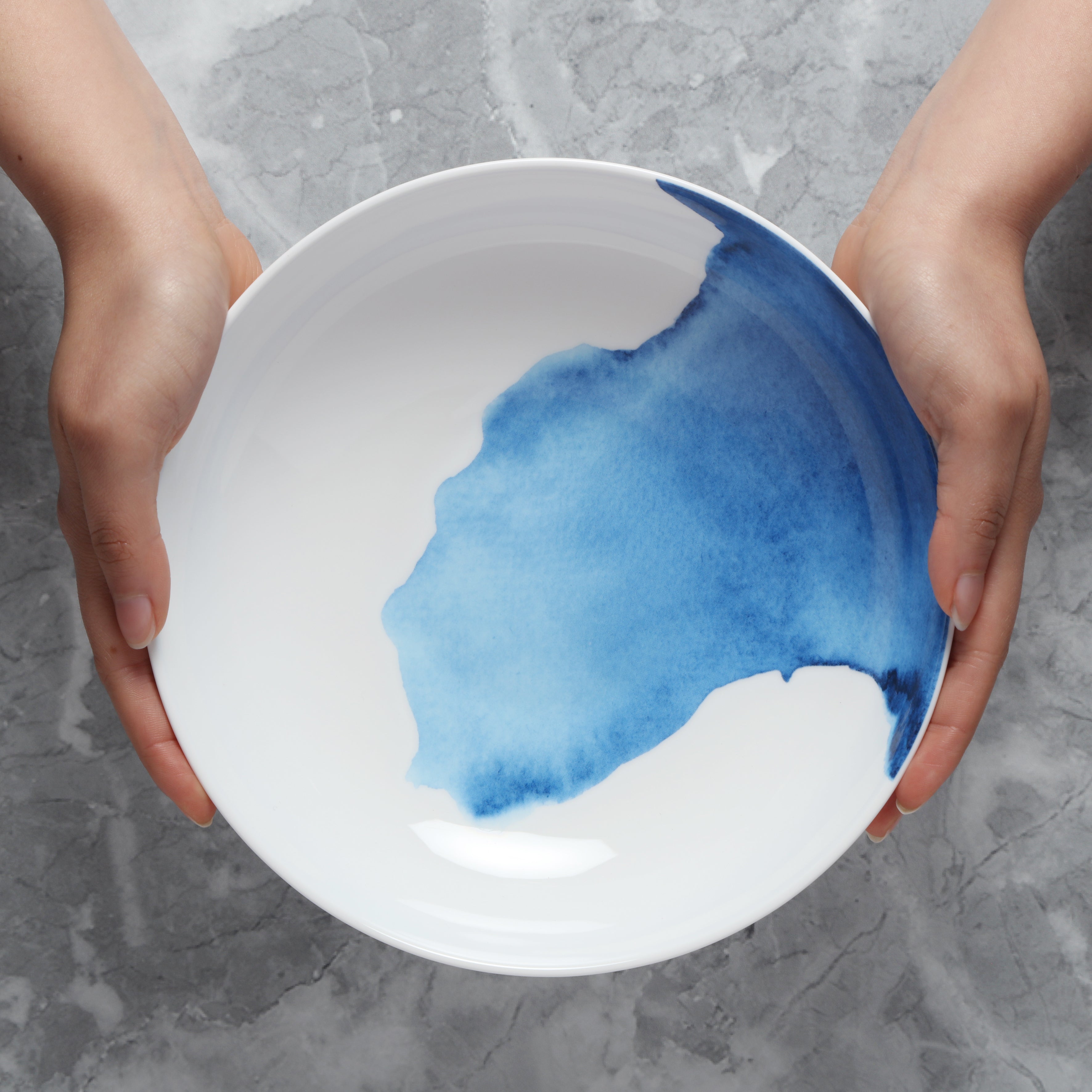 White supper bowl with blue watercolor design held by hands on a gray surface