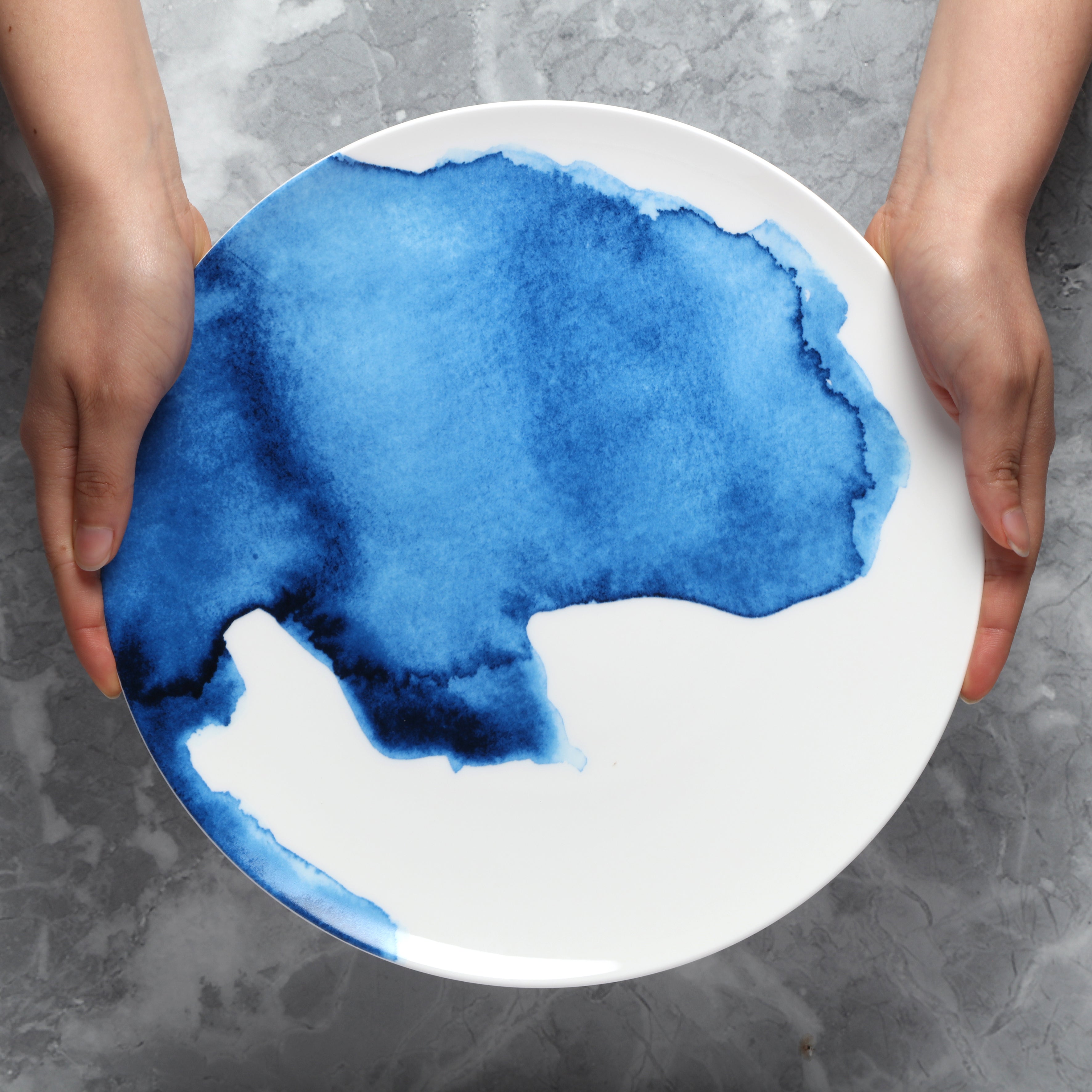 Person holding a white plate with blue watercolour design on a gray surface