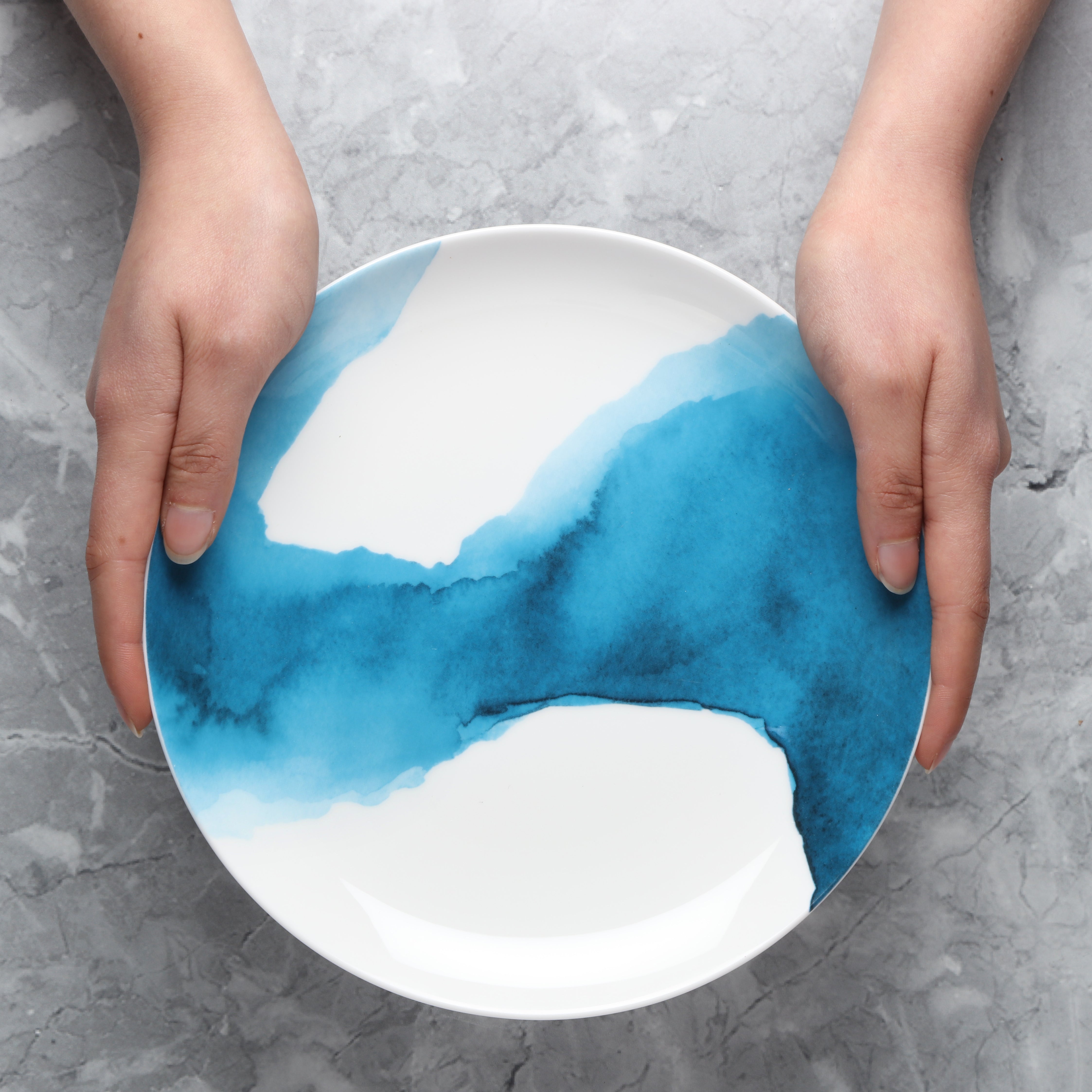 Person holding a ceramic side plate with blue and white watercolour design on a gray surface